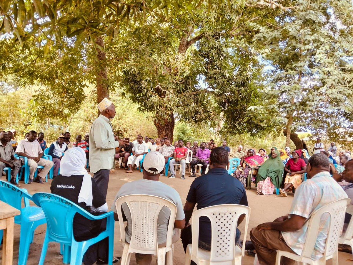 Community members seated in a circle beneath shade trees as an elder addresses the gathering.